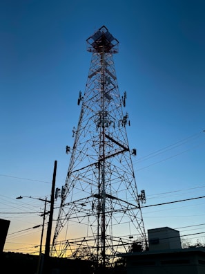 A tall telecommunications tower silhouetted against a clear blue sky during sunset, with various antennas attached at different heights. Power lines and utility poles are visible in the foreground, along with some buildings and trees at the base of the tower.