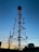 A tall telecommunications tower silhouetted against a clear blue sky during sunset, with various antennas attached at different heights. Power lines and utility poles are visible in the foreground, along with some buildings and trees at the base of the tower.