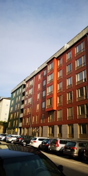 A row of modern apartment buildings with red and green facades, featuring multiple windows and a flat roof. Below them is a street lined with parked cars. The sky is clear and blue, suggesting a sunny day.