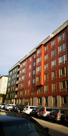 A row of modern apartment buildings with red and green facades, featuring multiple windows and a flat roof. Below them is a street lined with parked cars. The sky is clear and blue, suggesting a sunny day.