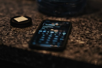 A smartphone lying on a textured stone surface displaying a blurred notification screen. Next to it, a lens cap with a handwritten label is visible. The background is softly focused, suggesting an intimate or thoughtful setting.