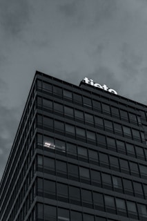 A modern multi-story building with a grid-like facade, displaying the word 'tieto' at the top. The overcast sky adds a moody atmosphere. The building's windows reflect the subdued light, with some illuminated from within.