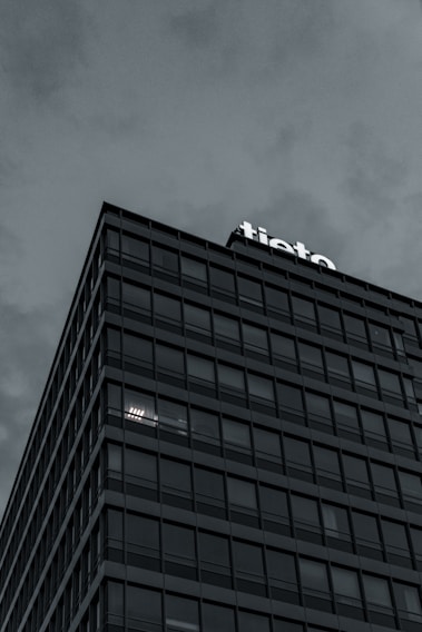 A modern multi-story building with a grid-like facade, displaying the word 'tieto' at the top. The overcast sky adds a moody atmosphere. The building's windows reflect the subdued light, with some illuminated from within.