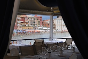 A beachfront view from inside a restaurant with several set tables and empty wine glasses. In the background, a lively beach with numerous people enjoying the water and colorful beach umbrellas can be seen. The backdrop features charming multicolored buildings typical of a coastal town.