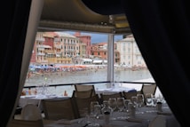 A beachfront view from inside a restaurant with several set tables and empty wine glasses. In the background, a lively beach with numerous people enjoying the water and colorful beach umbrellas can be seen. The backdrop features charming multicolored buildings typical of a coastal town.