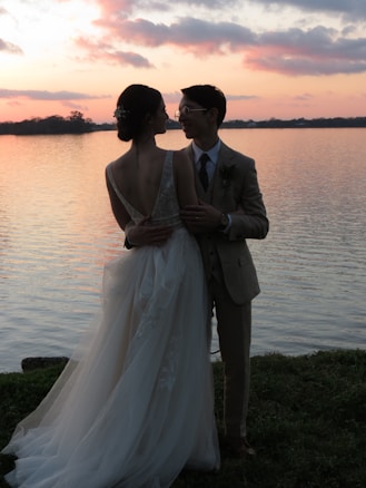 A couple stands by a serene lake during sunset. The sky is painted with hues of orange, pink, and purple, which reflect on the calm water. The woman wears an elegant white gown with a low back, and the man is dressed in a beige suit. They are gazing into each other's eyes, creating a romantic ambiance.