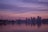 A city skyline is silhouetted against a colorful sunset, with a large body of water in the foreground reflecting the pastel hues of the sky. The iconic Space Needle is visible to the left, while the lit-up buildings of the downtown area create a striking contrast against the serene sky.