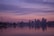 A city skyline is silhouetted against a colorful sunset, with a large body of water in the foreground reflecting the pastel hues of the sky. The iconic Space Needle is visible to the left, while the lit-up buildings of the downtown area create a striking contrast against the serene sky.