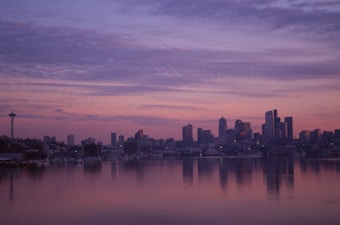 A city skyline is silhouetted against a colorful sunset, with a large body of water in the foreground reflecting the pastel hues of the sky. The iconic Space Needle is visible to the left, while the lit-up buildings of the downtown area create a striking contrast against the serene sky.