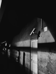 A black and white photo featuring a wall with a prominent security camera mounted on it. A metal door with horizontal slats is visible, casting stark shadows on the textured surface. The lighting creates dramatic contrasts between the bright light and the dark shadows.