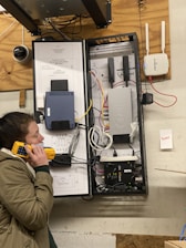 A person holding a yellow device stands next to an open telecommunications cabinet mounted on a wooden wall. The cabinet contains various networking and telecommunications equipment, including cables, routers, and a device with AT&T branding. A wall-mounted router with antennas is visible to the right.