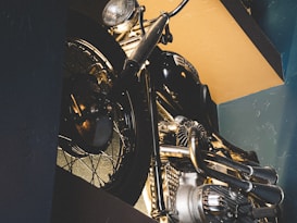 A vintage motorcycle is captured in a dimly lit setting, featuring intricate metallic details and a polished finish. The image highlights the wheel, engine, and handlebars, emphasizing the craftsmanship and design of the vehicle.