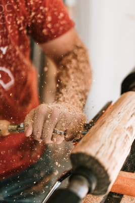 A person is using a lathe to shape a piece of wood. Wood shavings are scattered in the air, indicating an active woodworking process. The person is wearing a red shirt and holding the tool with a firm grip, focused on their work.