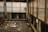 An abandoned industrial space features rusted metal beams and large windows with opaque glass obscured by grime. The floor is cluttered with litter, including papers, plastic, and other debris. A small patch of greenery protrudes into the frame near the windows, adding a touch of life to the otherwise neglected setting.