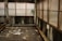 An abandoned industrial space features rusted metal beams and large windows with opaque glass obscured by grime. The floor is cluttered with litter, including papers, plastic, and other debris. A small patch of greenery protrudes into the frame near the windows, adding a touch of life to the otherwise neglected setting.