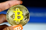 A person holds a physical representation of Bitcoin, with a large, prominent Bitcoin logo in yellow on a metallic gold coin. The background is a subtle gradient of blue and white, and the person's nails are painted orange with a Bitcoin symbol on one fingertip.