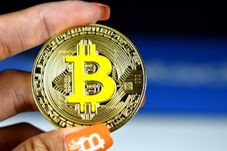 A person holds a physical representation of Bitcoin, with a large, prominent Bitcoin logo in yellow on a metallic gold coin. The background is a subtle gradient of blue and white, and the person's nails are painted orange with a Bitcoin symbol on one fingertip.