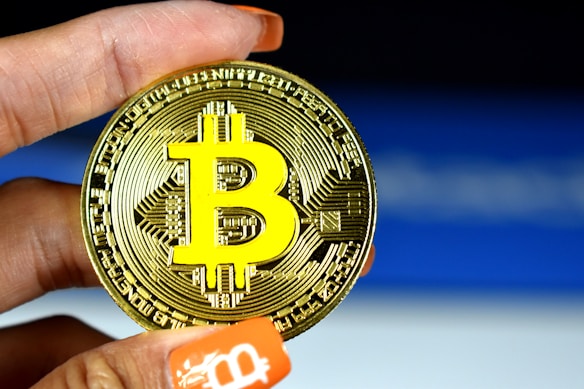A person holds a physical representation of Bitcoin, with a large, prominent Bitcoin logo in yellow on a metallic gold coin. The background is a subtle gradient of blue and white, and the person's nails are painted orange with a Bitcoin symbol on one fingertip.