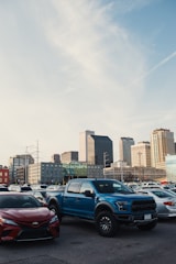 A parking lot is filled with vehicles, including a prominent blue pickup truck and a red sedan in the foreground. In the background, a city skyline with modern skyscrapers under a clear sky is visible.