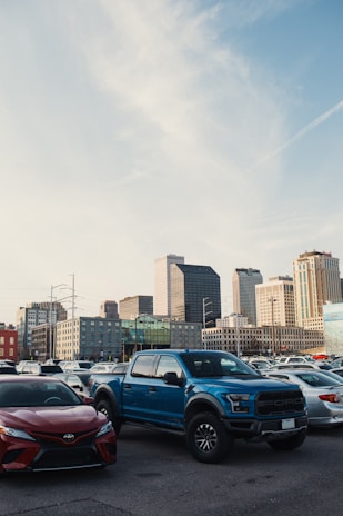 A parking lot is filled with vehicles, including a prominent blue pickup truck and a red sedan in the foreground. In the background, a city skyline with modern skyscrapers under a clear sky is visible.
