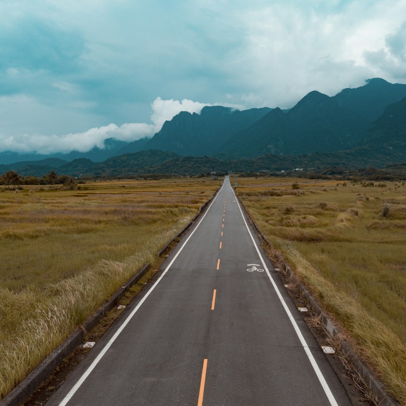 A straight two-lane road running between golden grassland towards green mountains under low clouds in Taitung, Taiwan