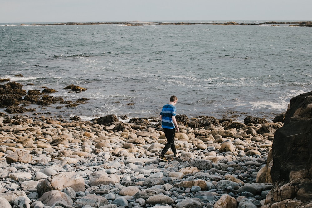 Chico con camisa azul y pantalones negros caminando en la orilla rocosa durante el día