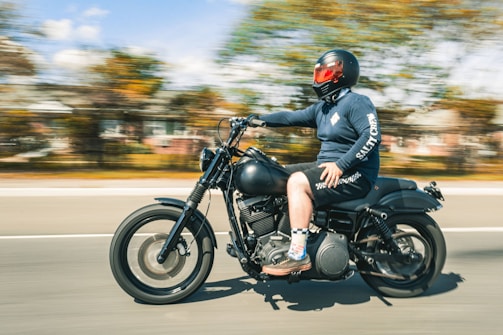 A person wearing protective gear, including a black motorcycle helmet with a red visor, rides a black motorcycle at speed. They are wearing a long-sleeved shirt, shorts, and colorful socks paired with loafers. The background shows a blurred view of trees and buildings, indicating motion.