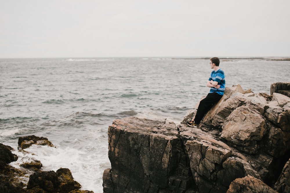 man in blue shirt sitting on rock formation near sea during daytime