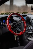 A car interior featuring a vibrant red and black steering wheel with a sleek design, an array of dashboard controls and gauges, a hanging air freshener, and a smartphone holder attached near the console. Keys are hanging from the steering wheel.