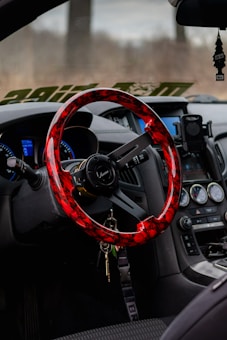A car interior featuring a vibrant red and black steering wheel with a sleek design, an array of dashboard controls and gauges, a hanging air freshener, and a smartphone holder attached near the console. Keys are hanging from the steering wheel.