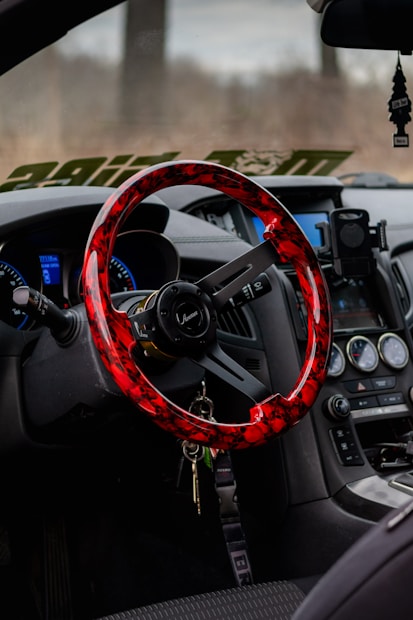 A car interior featuring a vibrant red and black steering wheel with a sleek design, an array of dashboard controls and gauges, a hanging air freshener, and a smartphone holder attached near the console. Keys are hanging from the steering wheel.