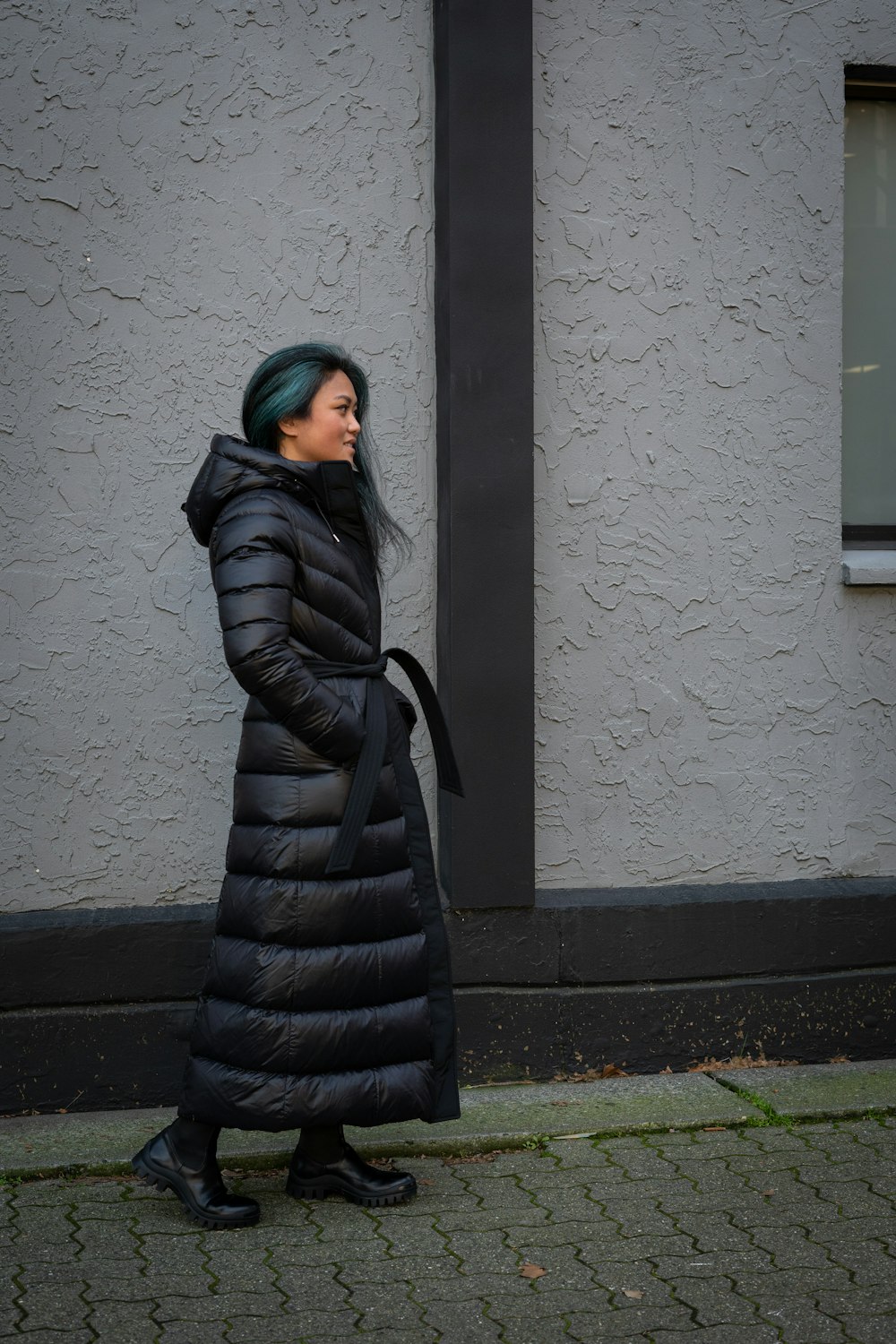 낮 동안 흰색 콘크리트 벽 옆에 서 있는 검은 거품 재킷을 입은 여자
