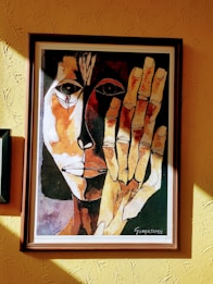A framed painting of an abstract face with contrasting dark and light tones. The artwork features bold, expressive linework with one half of the face in shadow and the other brightly lit. A large hand is prominently displayed in front of the face, matching the abstract style.