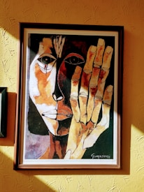 A framed painting of an abstract face with contrasting dark and light tones. The artwork features bold, expressive linework with one half of the face in shadow and the other brightly lit. A large hand is prominently displayed in front of the face, matching the abstract style.
