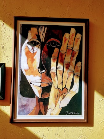 A framed painting of an abstract face with contrasting dark and light tones. The artwork features bold, expressive linework with one half of the face in shadow and the other brightly lit. A large hand is prominently displayed in front of the face, matching the abstract style.