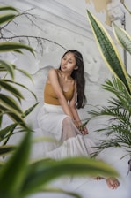 A woman with long dark hair sits peacefully surrounded by lush green indoor plants against a light-colored textured wall. She wears a sleeveless brown top and a flowing white skirt, and there are decorative branches visible on the wall.