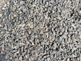 A densely packed pile of small, irregularly shaped gray and brown stones with various textures and shades. The stones appear rough and are closely arranged, suggesting they might be used for construction or landscaping purposes.