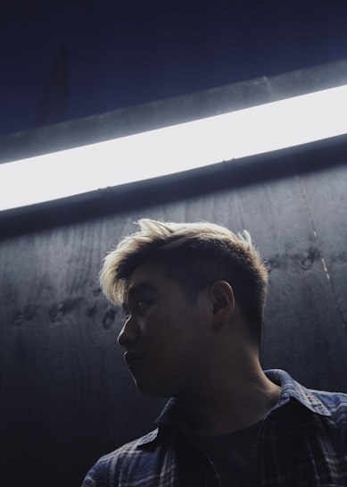 A person is seen in profile against a dark background, illuminated by a bright fluorescent light above them. The lighting creates a strong contrast, highlighting their face and hair while leaving much of their form in shadow. The setting appears to be urban, with an industrial or gritty feel.