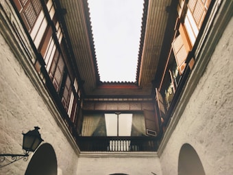 A courtyard surrounded by intricately designed balconies and wooden lattice windows. The architecture features a mix of white stone walls and dark wooden elements. A vintage lantern hangs from one side, contributing to a historical or classical ambiance. Natural light filters through the open roof, creating a serene atmosphere.