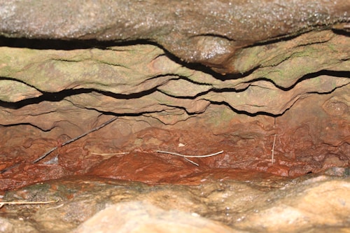 Layers of rock with varying shades of brown and green, displaying a textured and eroded surface. The rock appears to be formed naturally, with a rough and rugged appearance, suggesting an underground cave or a similar formation.
