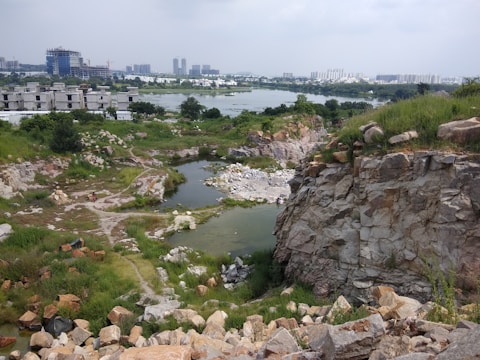 A landscape featuring a rocky terrain with patches of greenery and a small body of water. In the background, there are several construction buildings and a distant view of a city skyline with tall buildings. The scene is cloudy, casting a neutral light over the area.