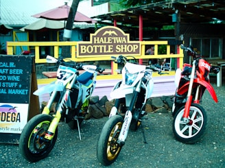 Three motorcycles are parked in front of a rustic shop with a sign reading 'Hale'iwa Bottle Shop'. The bikes display numbers and logos, with the central bike being predominantly white, the left one featuring a mix of blue and yellow, and the right one primarily red. A wooden sign advertising craft beer and wine is positioned to the left. The scene is set in an outdoor market area with a casual, laid-back atmosphere.