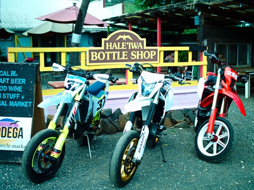 Three motorcycles are parked in front of a rustic shop with a sign reading 'Hale'iwa Bottle Shop'. The bikes display numbers and logos, with the central bike being predominantly white, the left one featuring a mix of blue and yellow, and the right one primarily red. A wooden sign advertising craft beer and wine is positioned to the left. The scene is set in an outdoor market area with a casual, laid-back atmosphere.