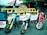 Three motorcycles are parked in front of a rustic shop with a sign reading 'Hale'iwa Bottle Shop'. The bikes display numbers and logos, with the central bike being predominantly white, the left one featuring a mix of blue and yellow, and the right one primarily red. A wooden sign advertising craft beer and wine is positioned to the left. The scene is set in an outdoor market area with a casual, laid-back atmosphere.