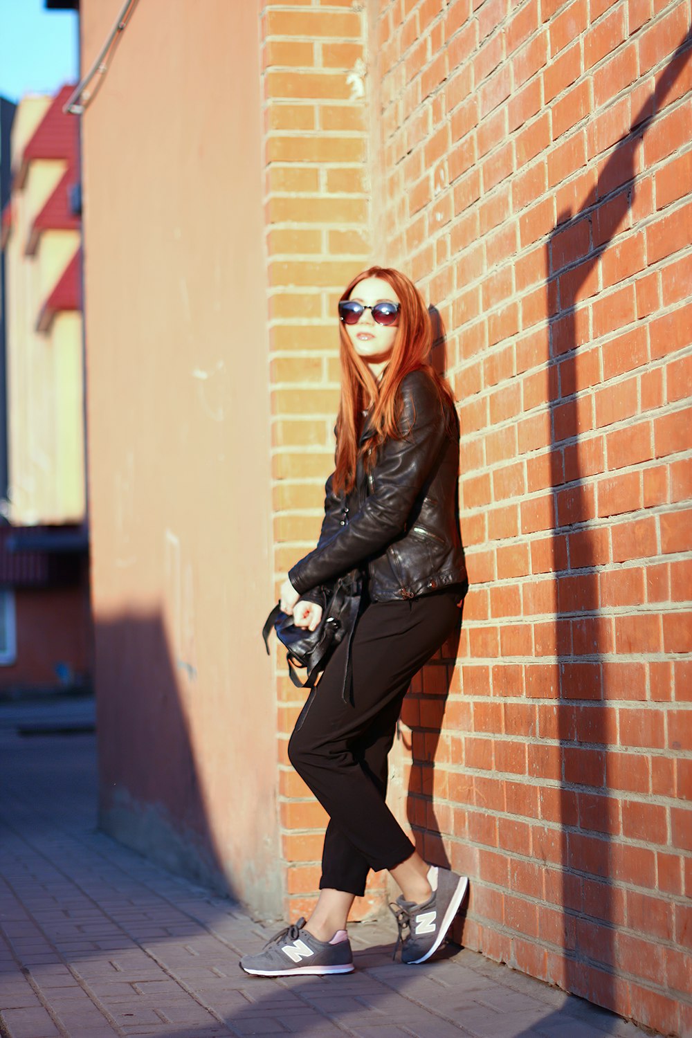 Femme en veste en cuir noir appuyée sur un mur de briques brunes pendant la journée