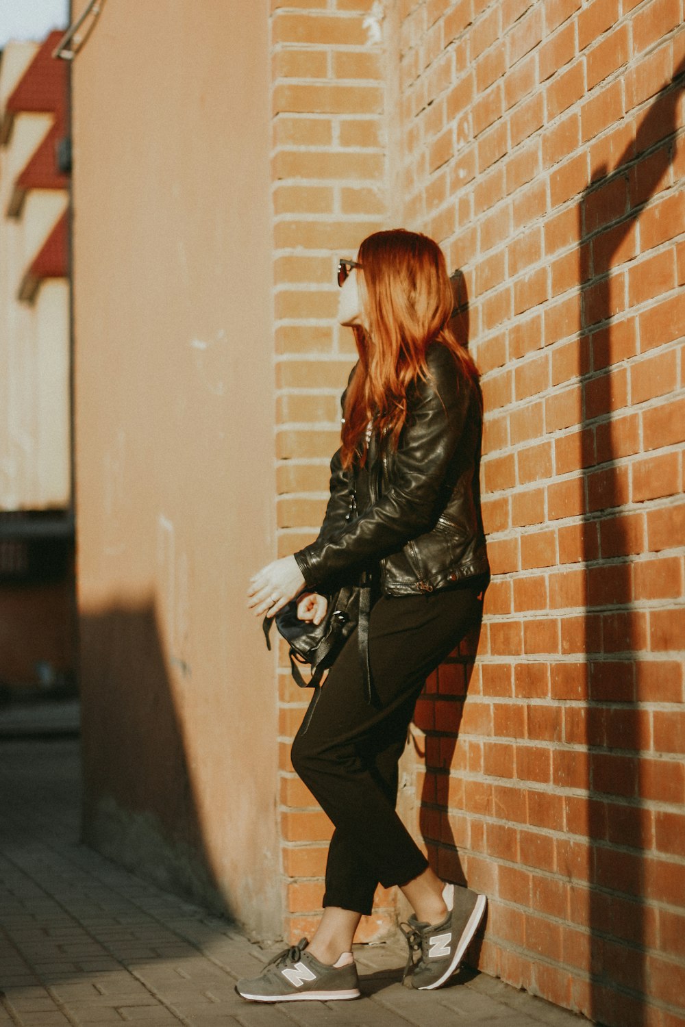 woman in black leather jacket and black pants leaning on brown brick wall during daytime