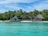 A picturesque tropical beach scene featuring two thatched-roof huts surrounded by lush palm trees. The huts are situated directly on the sandy shoreline, with a deck area that looks over the turquoise waters, giving off a serene and inviting atmosphere.