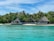 A picturesque tropical beach scene featuring two thatched-roof huts surrounded by lush palm trees. The huts are situated directly on the sandy shoreline, with a deck area that looks over the turquoise waters, giving off a serene and inviting atmosphere.