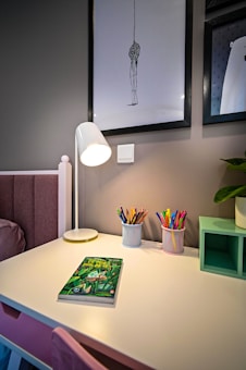 A brightly lit desk featuring a white lamp and containers filled with colorful pencils. There is a book with a green cover resting on the desk. The wall behind the desk has framed artwork, and a small shelf holding a plant. The scene is cozy and organized.