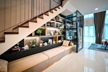 A modern interior space features an under-staircase shelving unit adorned with decorative items and indoor plants. The space includes a beige cushioned bench, a glass-encased lift, and a large window with sheer curtains allowing natural light. A cozy atmosphere is complemented by contemporary furniture and decor.